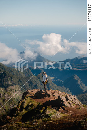 Hiker stands atop rocky heights viewing vast ocean and fluffy clouds Hiker stands atop rocky heights viewing vast ocean and fluffy clouds 135374317