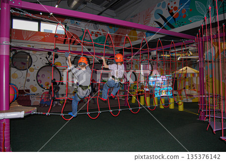 Kids navigating an indoor rope course at an adventure park. Children wearing helmets navigate a red rope obstacle at an indoor adventure park. Kids navigating an indoor rope course at an adventure park. Children wearing helmets navigate a red rope obstacle at an indoor adventure park. 135376142
