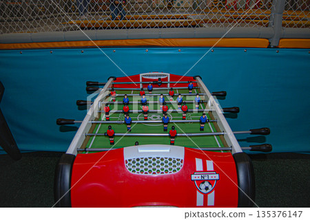 Red foosball table ready for a game. Red foosball table with a green playing field and plastic player figures arranged in traditional formation. Red foosball table ready for a game. Red foosball table with a green playing field and plastic player figures arranged in traditional formation. 135376147