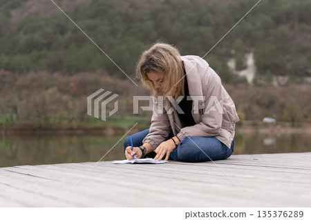 Woman writing notebook Young woman writing in notebook while sitting on wooden dock near forest lake planning future Woman writing notebook Young woman writing in notebook while sitting on wooden dock near forest lake planning future 135376289