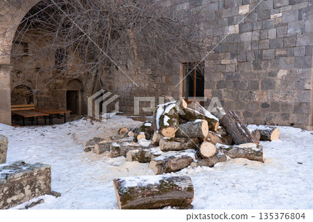 Stone courtyard of Tatev monastery with arched passage, bare tree, stacked firewood, snow on the ground, muted winter colors, quiet rustic atmosphere 135376804