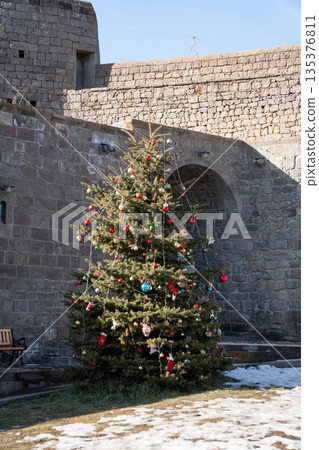Decorated Christmas tree stands in stone courtyard near historic monastery walls, evergreen branches, colorful ornaments, winter, calm festive mood 135376811