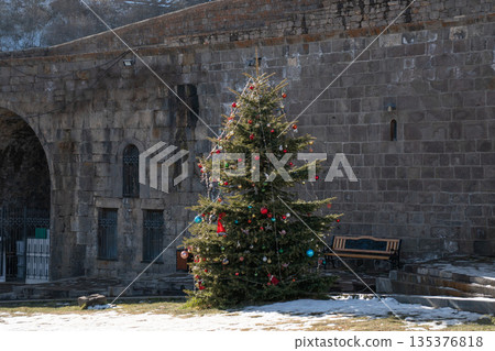 Decorated Christmas tree stands in stone courtyard near historic monastery walls, evergreen branches, colorful ornaments, winter, calm festive mood Decorated Christmas tree stands in stone courtyard near historic monastery walls, evergreen branches, colorful ornaments, winter, calm festive mood 135376818
