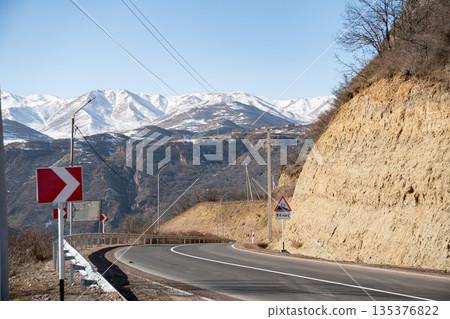 Winding mountain road with sharp turn signs cutting through a rocky hillside, with snow-covered peaks and deep valley in the background under blue sky 135376822
