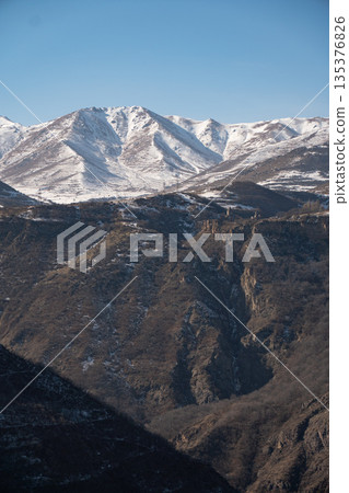 Snow-covered mountain ridges, deep gorge landscape in Armenia with Tatev Monastery visible in the distance, clear winter sky, dramatic natural relief 135376826