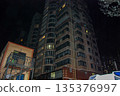 Tall residential building illuminated at night in an urban setting. The structure features multiple floors with rows of windows, some of which are lit, showcasing a modern architectural design with a  135376997
