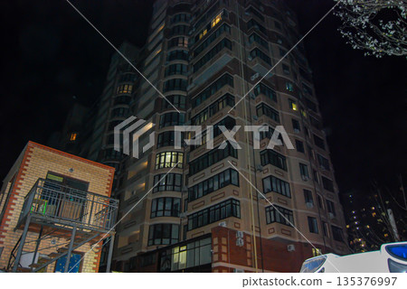 Tall residential building illuminated at night in an urban setting. The structure features multiple floors with rows of windows, some of which are lit, showcasing a modern architectural design with a Tall residential building illuminated at night in an urban setting. The structure features multiple floors with rows of windows, some of which are lit, showcasing a modern architectural design with a 135376997