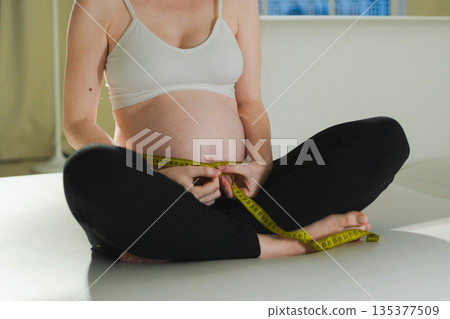 Pregnancy motherhood. Pregnant woman measuring big belly. Girl measuring her belly with tape to keep track of her fetus development. Woman enjoying pregnancy. Maternity tenderness parenthood new life Pregnancy motherhood. Pregnant woman measuring big belly. Girl measuring her belly with tape to keep track of her fetus development. Woman enjoying pregnancy. Maternity tenderness parenthood new life 135377509