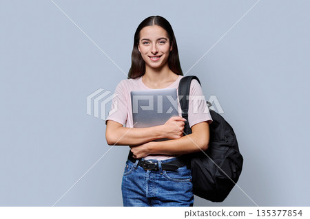 Portrait of beautiful female teenage student with backpack, laptop on gray background Portrait of beautiful female teenage student with backpack, laptop on gray background 135377854