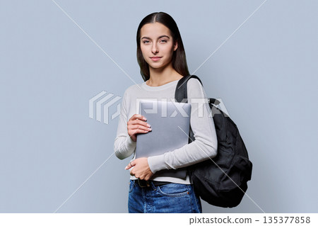 Portrait of beautiful female teenage student with backpack, laptop on gray background Portrait of beautiful female teenage student with backpack, laptop on gray background 135377858