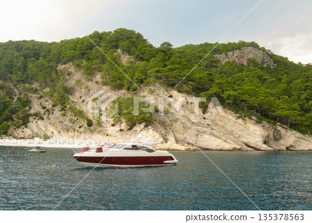 Luxury yacht anchored by rocky coast with pine hills and beach cabanas horizontal 135378563