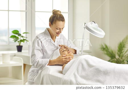 Woman cosmetologist checking client skin condition under lamp in beauty salon. Woman cosmetologist checking client skin condition under lamp in beauty salon. 135379632