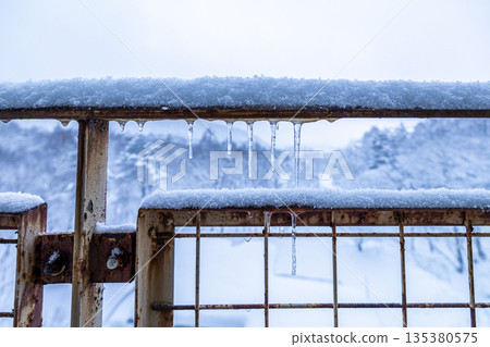 冬季景象:鏽跡斑斑的金屬柵欄上覆蓋著積雪和冰柱。 冬季景象:鏽跡斑斑的金屬柵欄上覆蓋著積雪和冰柱。 135380575