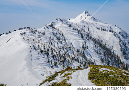 登山者沿著白雪皚皚的山脊行走 | 晴朗的冬季山景 登山者沿著白雪皚皚的山脊行走 | 晴朗的冬季山景 135380593