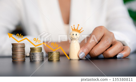 Businesswoman hand holding smiling wooden king doll with decreasing coin stacks and percentage graph for cost reduction concept. 135380811