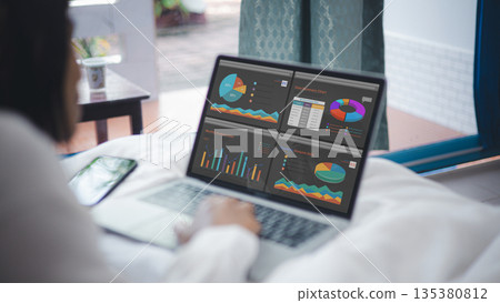 Businesswoman working on laptop in bed showing financial analysis dashboard. Concept of remote work, digital marketing statistics, data visualization, and business strategy success. 135380812