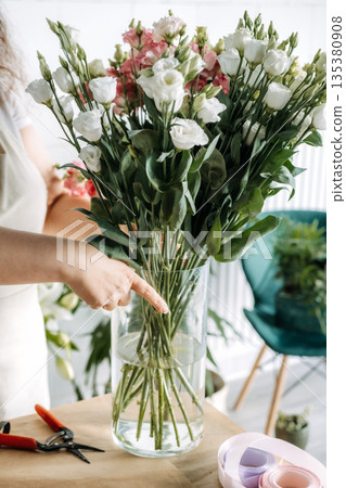 Person places stems of Lisianthus flowers into glass vase on craft paper covered table. Florist education, DIY botanical workshop, flower arranging class. Person places stems of Lisianthus flowers into glass vase on craft paper covered table. Florist education, DIY botanical workshop, flower arranging class. 135380908