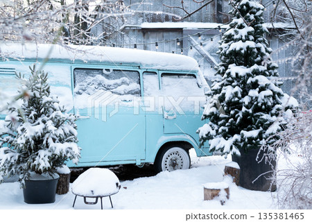 Vintage blue camper van parked in a snowy winter yard with snow-covered pine trees and decorative string lights. Vintage blue camper van parked in a snowy winter yard with snow-covered pine trees and decorative string lights. 135381465