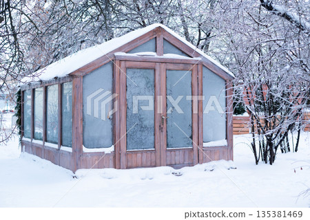 A wooden garden greenhouse covered in snow and frost, surrounded by icy winter trees in a backyard A wooden garden greenhouse covered in snow and frost, surrounded by icy winter trees in a backyard 135381469