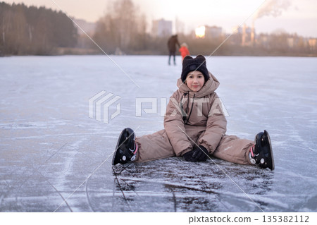 A happy girl wearing ice skates sits on the surface of a frozen lake while her father and sister skate 135382112