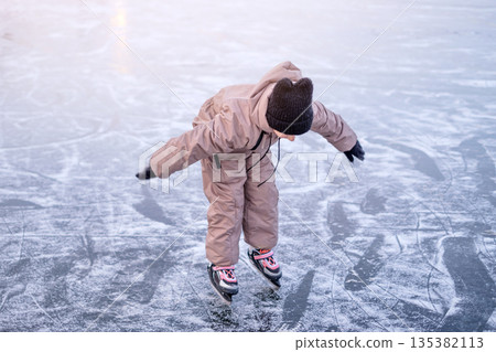 A child tries to keep balance while learning to skate on the ice rink of a frozen lake. A child tries to keep balance while learning to skate on the ice rink of a frozen lake. 135382113