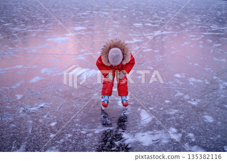 A tired child while skating. Reflection of the red setting sun on the ice of the frozen sun in the late evening 135382116