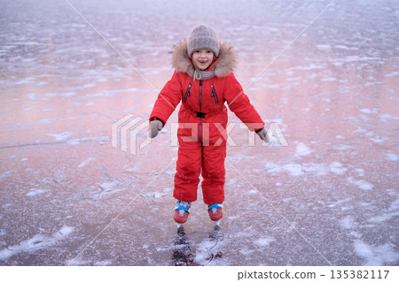 A laughing child confidently stands on ice skates, skating on a cold winter day on the ice of a frozen lake 135382117