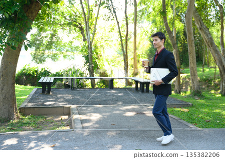 A man walks through a green park holding a laptop and a coffee cup on a bright day 135382206