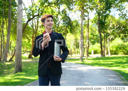 A man walks through a green park holding a laptop and a coffee cup on a bright day 135382229