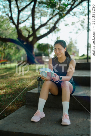 Smiling woman resting and listening to music during outdoor workout break in park Smiling woman resting and listening to music during outdoor workout break in park 135382250