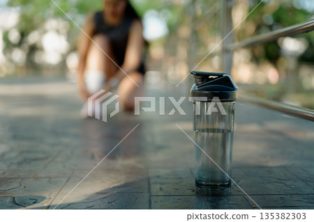 Water bottle in focus as female runner adjusts shoe during outdoor workout break in park Water bottle in focus as female runner adjusts shoe during outdoor workout break in park 135382303