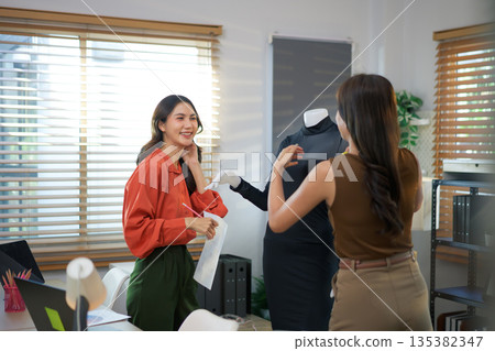 Two women measuring a dress on a mannequin in a home fashion studio, smiling while planning design details, creative teamwork concept 135382347