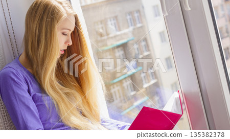 Woman sitting on window sill reading book at home Woman sitting on window sill reading book at home 135382378