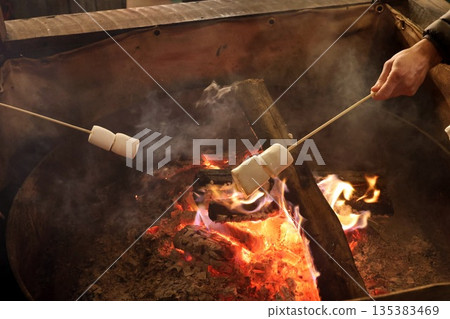 Hands roasting marshmallows on wooden skewers over a glowing campfire. Hands roasting marshmallows on wooden skewers over a glowing campfire. 135383469