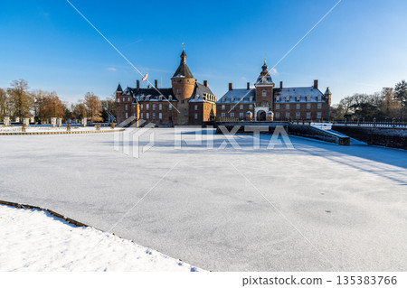 Snow covered castle with red brick towers and spire rises above frozen moat under clear blue sky, evoking winter grandeur. Snow covered castle with red brick towers and spire rises above frozen moat under clear blue sky, evoking winter grandeur. 135383766