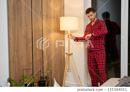 Man in red pajamas standing by floor lamp and checking wristwatch in living room Man in red pajamas standing by floor lamp and checking wristwatch in living room 135384051