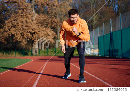 Man in orange sports jacket starting sprint on outdoor running track in stadium 135384053