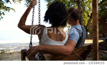 Romantic couple ride on swing together at sea beach. Happy lovers enjoy to spending time together at resort. Carefree pair enjoying rest near ocean at sunny day. Concept of summer vacation or holiday 135384133