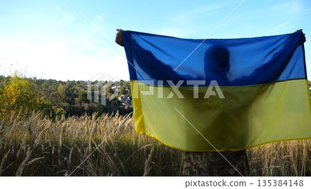 Military man in uniform stands with raised over head flag of Ukraine at countryside. Male ukrainian army soldier with lifted national banner in honor of victory against russian aggression. End of war Military man in uniform stands with raised over head flag of Ukraine at countryside. Male ukrainian army soldier with lifted national banner in honor of victory against russian aggression. End of war 135384148
