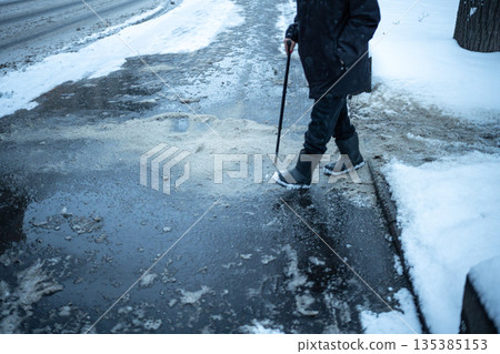 Person with cane carefully walking on pavement coated with technical salt and wet snow in winter. 135385153