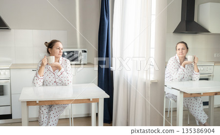 Woman sits at kitchen table in her pajamas, enjoying quiet morning with cup of coffee at home. Slow life, relaxed lifestyle Woman sits at kitchen table in her pajamas, enjoying quiet morning with cup of coffee at home. Slow life, relaxed lifestyle 135386904