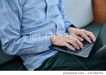 A man in his 60s sitting on a sofa and operating a laptop 135388741