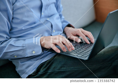 一位六十多歲的男子坐在沙發上操作筆記型電腦。 135388742