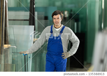 Man glazer employee in blue overall works in glass processing shop Man glazer employee in blue overall works in glass processing shop 135388913