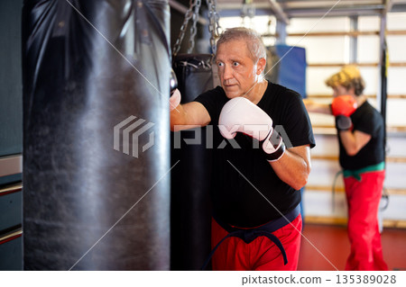 Elderly man trains in the gym - practicing punches on punching bag Elderly man trains in the gym - practicing punches on punching bag 135389028