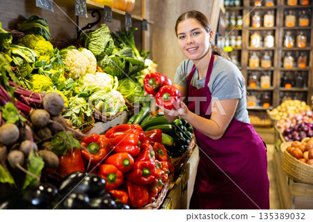 Female seller holding red bell pepper standing in fruit and vegetable section of supermarket Female seller holding red bell pepper standing in fruit and vegetable section of supermarket 135389032