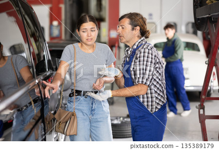Worried girl points to scratches on car after accident to auto mechanic at auto repair shop Worried girl points to scratches on car after accident to auto mechanic at auto repair shop 135389179