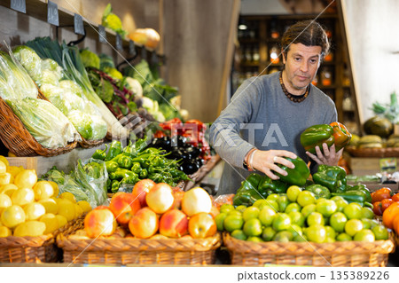 Man diligently picking out green bell pepper to buy in vegetable aisle of supermarket Man diligently picking out green bell pepper to buy in vegetable aisle of supermarket 135389226