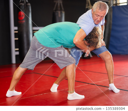 Mature man and adult man practice self-defense techniques at gym 135389304