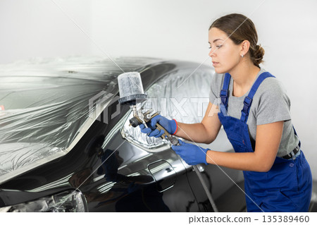 Car painting and automobile repair service. Woman in blue overalls paints car with airbrush pulverizer in paint chamber Car painting and automobile repair service. Woman in blue overalls paints car with airbrush pulverizer in paint chamber 135389460